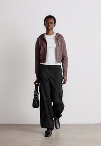 Brunet avskåret hettegenser, hvit skjorte, sorte cargo-bukser og svarte sko. Modellen holder en liten svart veske, stående mot en ensfarget bakgrunn.