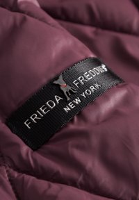 Bordeauxkleurige pufferstof met een gladde textuur, voorzien van een zwart etiket met "FRIEDA & FREDDIE NEW YORK" en een grijze hondenicoon.