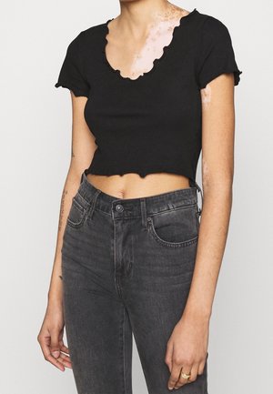 Person trägt ein schwarzes, kurzes, bauchfreies Shirt mit kurzen Ärmeln und hochtaillierte, ausgewaschene schwarze Jeans mit einem goldenen Ring an der linken Hand.