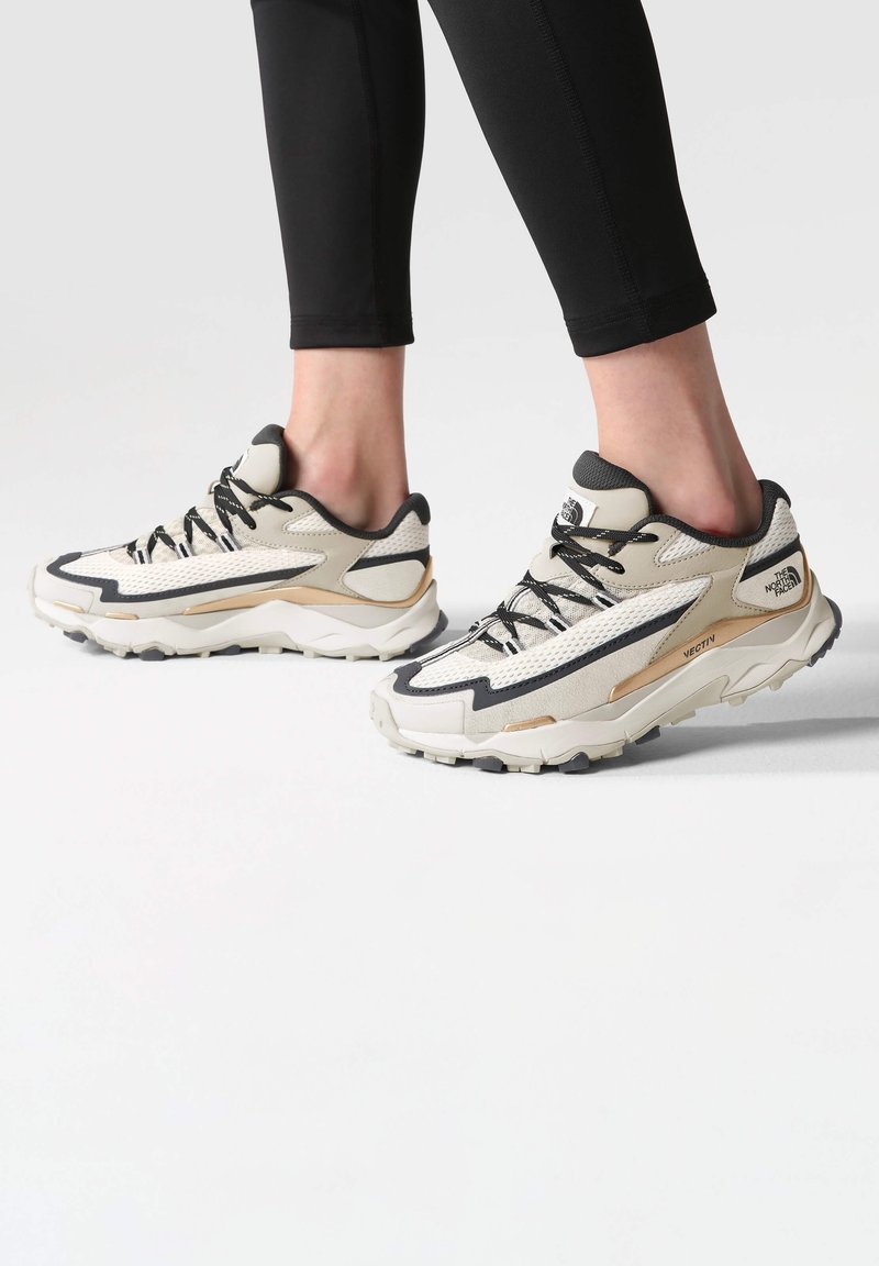 Zapatillas deportivas beige y negras con suelas resistentes, usadas por una persona en leggings negros que está de pie sobre una superficie blanca.