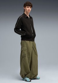 Zwarte ritsjack met witte piping, gecombineerd met losse olijfgroene cargobroek en tealkleurige sneakers met zilveren accenten. Soepele stofstructuren.