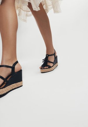 Sandales à talons noirs avec des brides de cheville ajustables, ornées de franges et d'une plateforme en jute tissé. Matière en cuir souple.