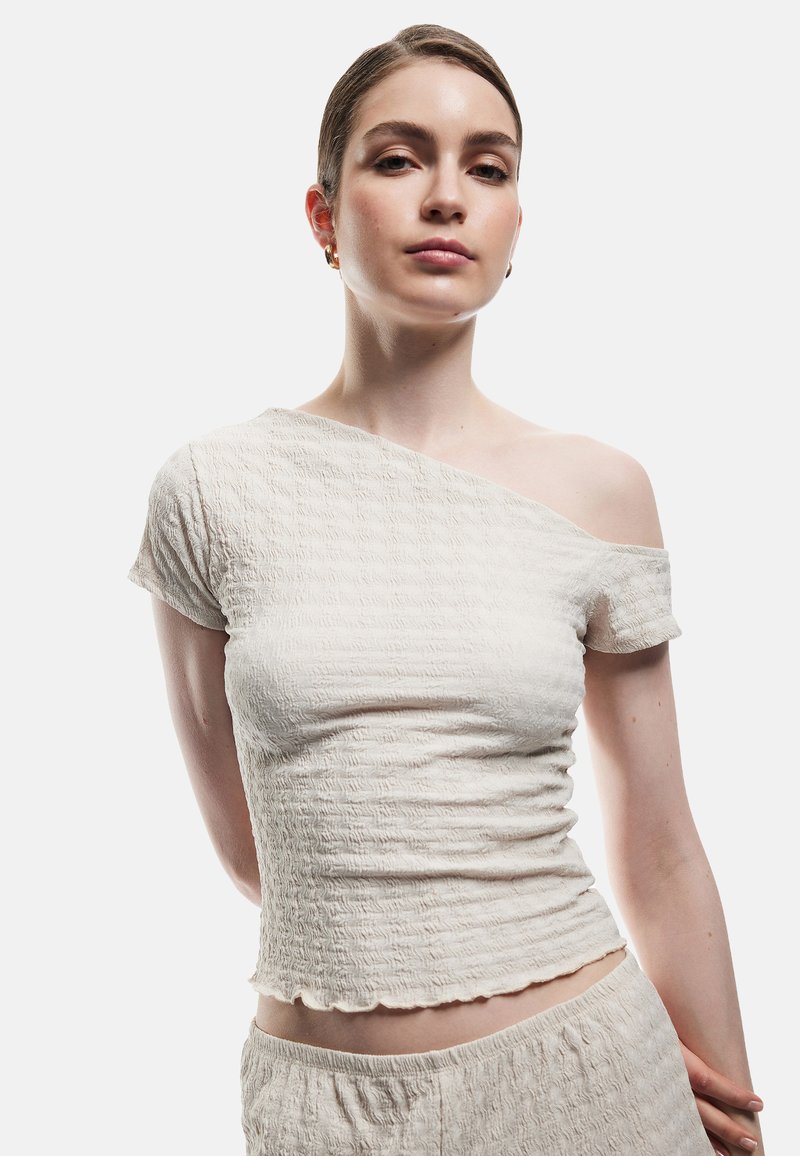 Off-shoulder cremetop med tekstureret stof, korte ærmer og bølget kant, parret med matchende bund. Glat, let materiale.