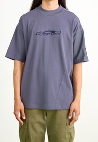 Camiseta de manga corta, de corte holgado, en algodón gris oscuro con un gráfico sutil de un coche de carreras en azul marino. Presenta un escote alto y un diseño de panel lateral.