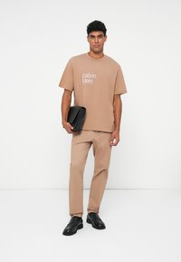 Rudas medvilninis marškinėlis su balta "calvin klein" logotipu, derinamas su atitinkančiomis kelnių ir juodais odiniais basutėmis, laiko juodą maišą.