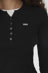 Primo piano di una persona che indossa una maglia nera a maniche lunghe a coste con una maglietta bianca sotto, bottoni neri e un piccolo logo bianco ricamato sul petto.