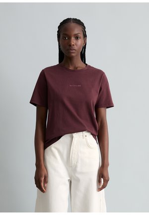 Frau, die vor einem einfarbigen Hintergrund steht, trägt ein kastanienbraunes T-Shirt mit kurzen Ärmeln und weiße, hoch taillierte Hosen, blickt geradeaus.