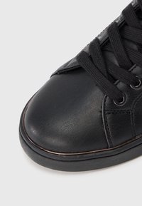 Primo piano di una punta di sneaker nera in pelle con lacci neri e occhielli metallici su uno sfondo grigio.