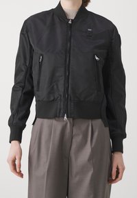 Černá bombera s předním zipem, pružnými manžetami, dvěma předními zipovými kapsami a malou nášivkou na levém horním rohu, vyrobena z hladkého materiálu.