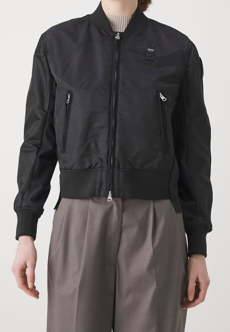 Černá bombera s předním zipem, pružnými manžetami, dvěma předními zipovými kapsami a malou nášivkou na levém horním rohu, vyrobena z hladkého materiálu.