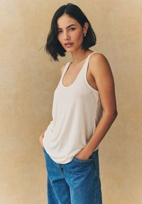 Débardeur blanc à côtes avec un col rond, associé à un jean bleu en denim ample. Sur fond texturé neutre.