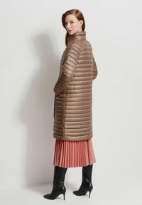 Brązowy puchowy płaszcz z pionowym pikowaniem, wysoka stójka i boczne kieszenie, w połączeniu z różową spódnicą plisowaną i czarnymi botkami na obcasie.