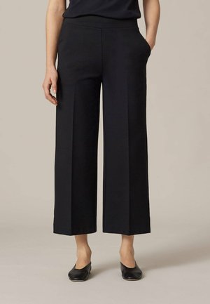 Zwarte wijde broek met een gladde textuur, voorzien van zijzakken en een getailleerde pasvorm. Afgewerkt met een nette zoom bij de enkel.