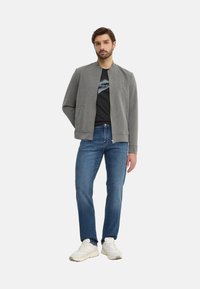 Chaqueta bomber gris con cremallera frontal, camiseta negra, vaqueros azules y zapatillas blancas. La chaqueta tiene un cuello redondo y bolsillos laterales.