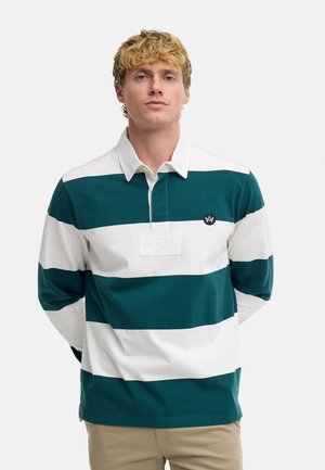 Junger Mann mit lockigem blondem Haar, trägt ein langärmeliges grün-weiß breit gestreiftes Rugbyshirt und beige Hose, steht vor einfachem Hintergrund.