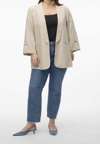 Nainen, jolla on päällään beige avointa etuosaa oleva bleiseri, musta paita, siniset suorat farkut frayed-helmalla, siniset matalakorkoiset slingback-kengät ja kukkakaulaketju.