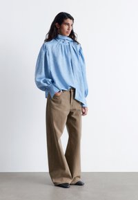 Himmelblaue, strukturierte Bluse mit hohem Kragen und puffartigen Ärmeln, kombiniert mit lockeren, beigefarbenen Hosen und schwarzen Schuhen. Schlichter weißer Hintergrund.