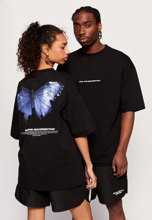 Zwei Models tragen schwarze Streetwear; Frau zeigt eine große blaue Schmetterlingsgrafik auf dem Rücken des Shirts, Mann schaut nach vorne mit weißem Text auf der Brust.