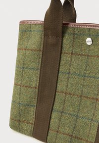 Πράσινη τσάντα από μάλλινο ύφασμα με καφέ κάθετους χειρολαβές, δερμάτινες λεπτομέρειες και κλείσιμο με μεταλλικό κουμπί που κουμπώνει, εμπνευσμένο από το λογότυπο.