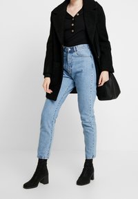 Manteau texturé noir, chemise noire à boutons, jean taille haute bleu clair et bottes noires à talons ; le mannequin tient un sac noir.