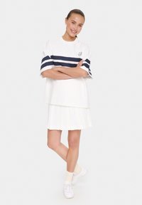 Camiseta blanca con rayas horizontales azul marino y logo; falda blanca con plisados; zapatillas blancas con calcetines claros. Look atlético casual.