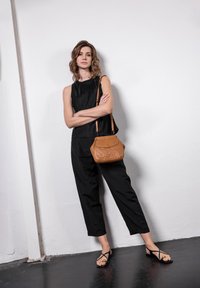 Top senza maniche nero e pantaloni neri larghi abbinati a sandali neri con cinturini. Una piccola borsa in pelle marrone pende da una tracolla.