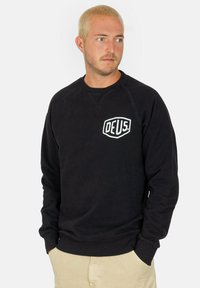 Sudadera negra con cuello y puños acanalados, que presenta un parche de logo blanco en el pecho. Textura suave con mangas largas y un ajuste holgado.