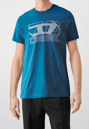 Homme portant un t-shirt Diesel bleu avec un logo rayé blanc et bleu, associé à un pantalon noir sur un fond uni.