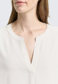 Blusa bianca con una texture morbida, scollatura a V e una vestibilità rilassata. Il tessuto presenta una finitura liscia con dettagli di cucitura sottili.
