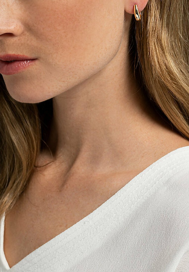 Guldörhänge i hoop-stil, slät yta, liten storlek, vilande på örsnibben. Närbild av en nacke, bärande en vit, texturerad klänning med V-hals.