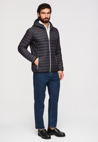 Homme debout, main dans la poche, portant une veste matelassée noire, une chemise blanche, un jean bleu et des chaussures noires sur un fond uni.