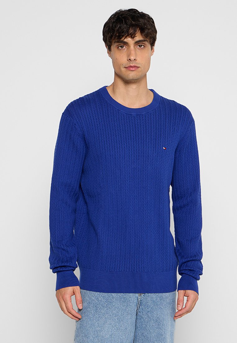 Kobaltblauwe gebreide trui met een textuur van kabelpatroon, ronde halslijn en ribgebreide manchetten en zoom. Klein logo-accent op de borst.