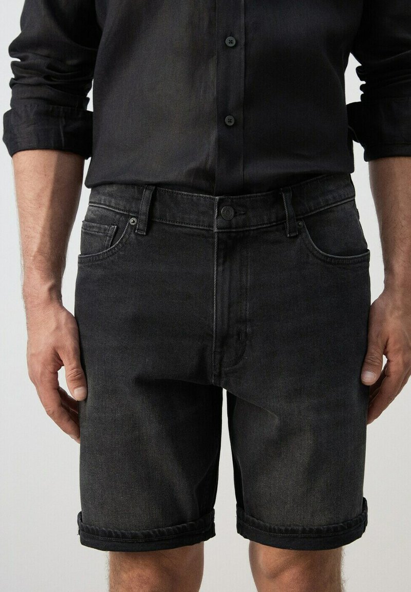 Homme portant un short en denim noir et une chemise noire à manches longues retroussées, debout les bras détendus le long du corps.