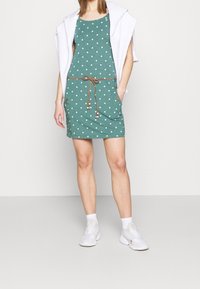 Robe verte à pois avec un design ajusté, taille coulissante et poches latérales ; associée à un sweat à capuche blanc et des baskets blanches.