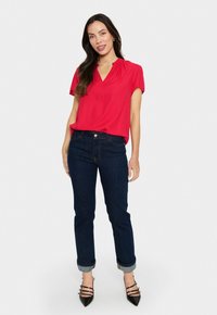 Blusa rossa a maniche corte con scollatura a V e tessuto morbido, abbinata a jeans blu scuro con risvolti e scarpe con tacco nere con multiple cinghie.