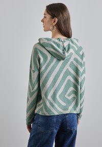 Sweat à capuche vert menthe avec un motif abstrait beige. Tissu doux avec une coupe décontractée, des épaules tombantes et des poignets côtelés.