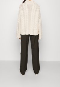 Kremowy sweter z grubym splotem i wysokim kołnierzem, noszony z ciemnymi, szerokimi spodniami. Sweter ma teksturowane wzory i luźny krój.