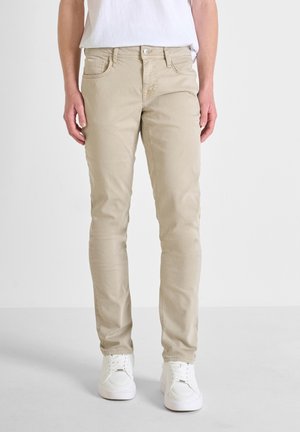 Vaqueros beige de corte slim, fabricados en una mezcla de algodón. Presentan bolsillos frontales, cierre de botón y detalles de costura sutiles. Combinados con zapatillas blancas.