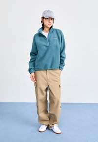 Pullover en polaire teal avec un zip quart et un rabat à boutons, associé à un pantalon cargo beige. Accessoirisé par une casquette rayée et des chaussures blanches.