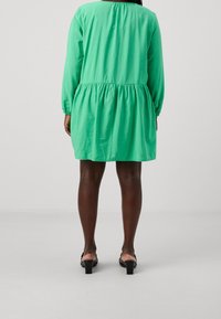 Grünes Langarmkleid mit geraffter Taille, runder Ausschnitt und elastischen Bündchen. Kombiniert mit schwarzen Sandalen mit Absatz.