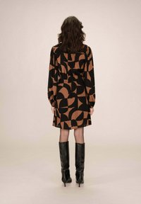 Robe à motifs noirs et bruns avec manches longues, taille cintrée et jupe qui arrive à mi-thigh, associée à des bottes noires montantes.