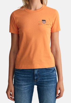 Mujer que lleva una camiseta naranja de manga corta con un pequeño logo de escudo bordado y texto en el pecho izquierdo, combinada con jeans azules.