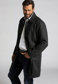 Abrigo de lana gris oscuro con cuello, cierre frontal de botones y bolsillos laterales. Lleva puesto una camisa blanca y jeans oscuros.