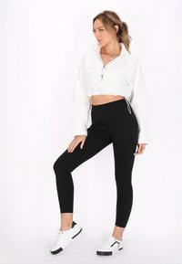 Felpa bianca corta con zip e vita con cordino, abbinata a leggings neri. Le calzature presentano una base bianca con accenti neri.