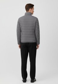 Grå puffjacka med horisontell quiltning, hög krage och avsmalnad passform. Bärs över svarta byxor. Slät textur, lättviktsdesign.