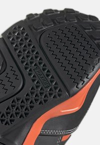 Semelle de chaussure en caoutchouc noir avec un motif texturé. Comprend des crampons triangulaires, des rainures en zigzag et un accent orange le long du côté.