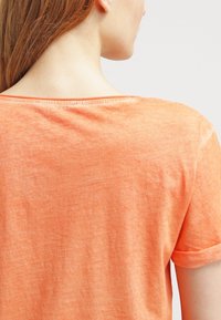 Žena s dlouhými hnědými vlasy v lehkém oranžovém tričku s krátkým rukávem, zobrazená zezadu od ramen až po hlavu na jednobarevném pozadí.