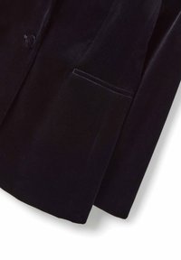 Dettaglio delle maniche e delle tasche di un blazer in velluto nero con un singolo bottone rivestito in tessuto sul davanti.