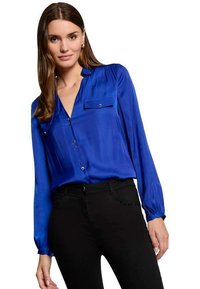 Blusa in satin blu con maniche lunghe e chiusura a bottoni. Presenta una tasca sul petto e una texture liscia e lucida. Abbinata a pantaloni neri.