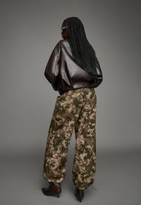 Bruine leren oversized jas, camouflagepatroon wijde broek in groen en beige, en bruine enkelhakken.
